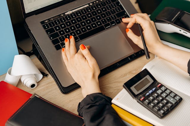 Woman typing on a laptop with a notepad and calculator between her arms